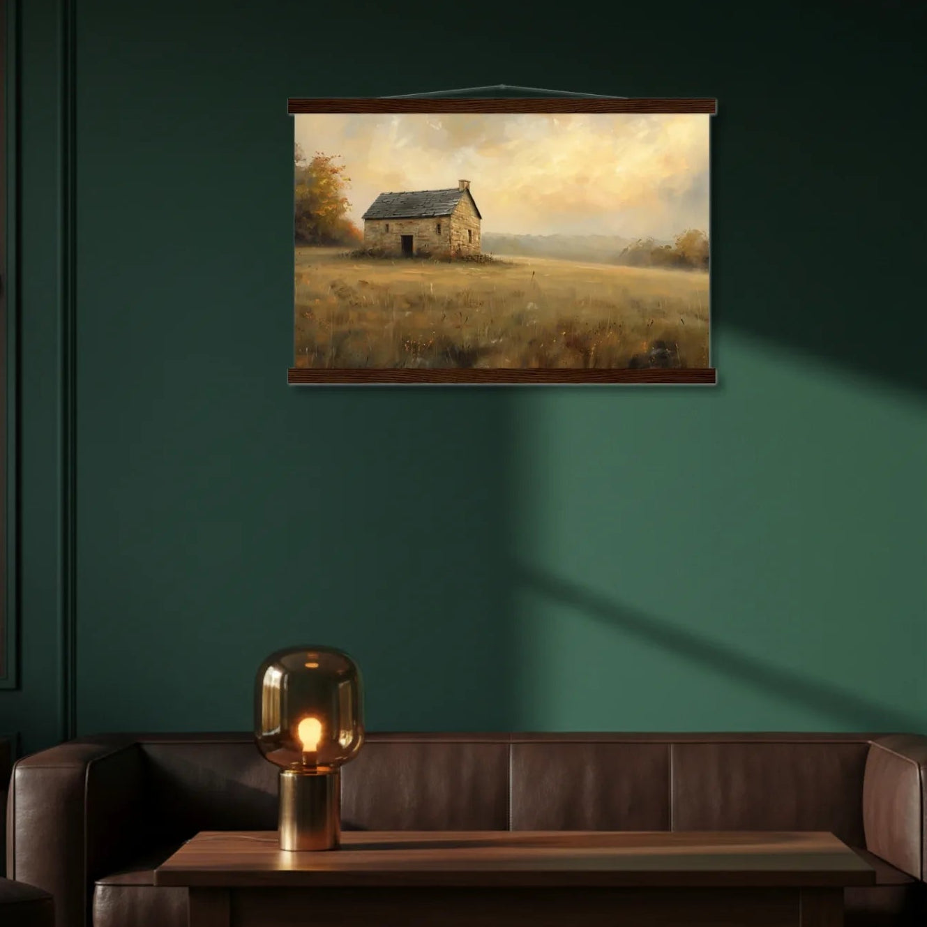 Framed painting of a small house in a field on a green wall above a brown leather sofa with a table and lamp.